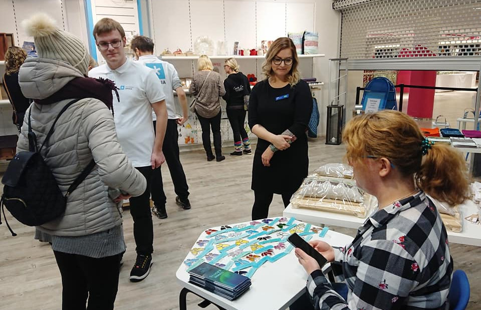 Ihmisiä ostoksilla puodissa, pöydän takana käsitöiden esittelijä.