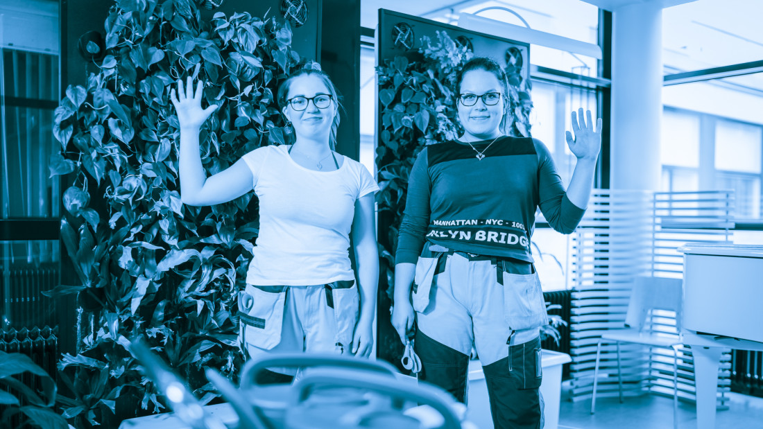 Kaksi nuorta naista on nostanut käden moikkaukseen, päällään heillä t-paidat ja työhousut. Kauttaaltaan sinisävyinen kuva.