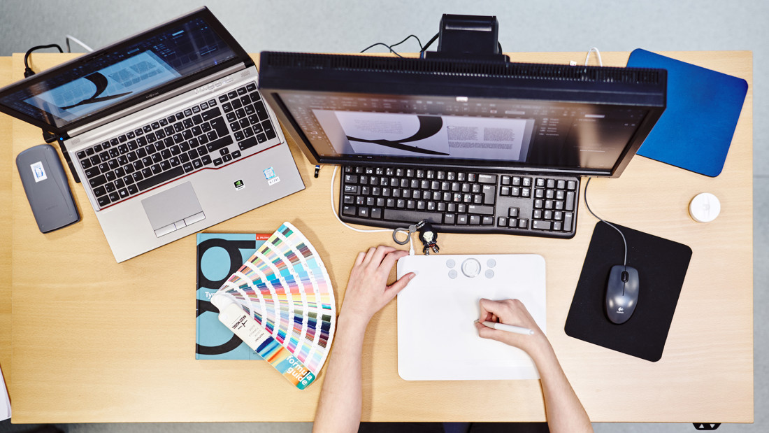 Ylhäältä kuvattuna kädet käyttävät piirtopäytää, edessään näyttö ja läppäri, vieressä väriviuhka.