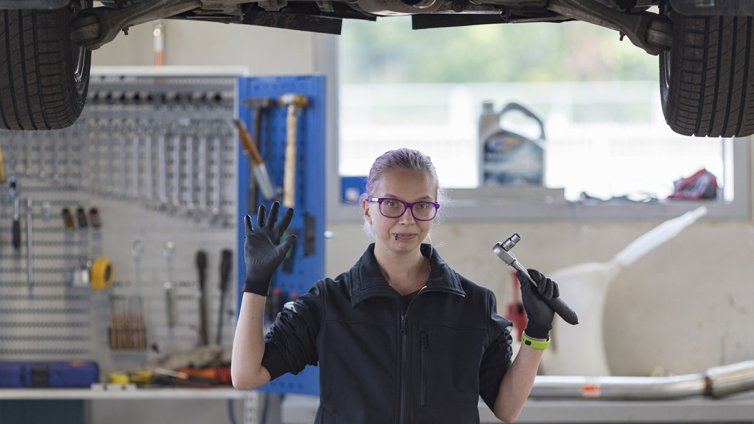 Ajoneuvoasentaja-opiskelija seisoo korkealle nostetun auton alla työkalu kädessään ja moikkaa.