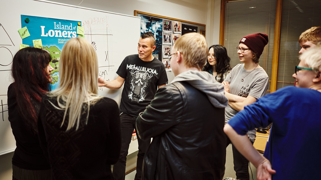 Opettaja ja nuoret seisovat ilmoitustaulun edessä ringissä ja katsovat kiinnostuneina jotain.