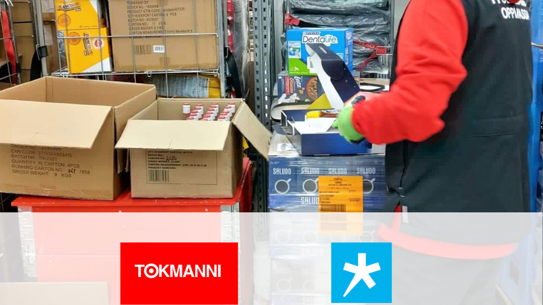 Lähikuva varastosta, jossa rullakko, laatikoita ja osittain näkyvä henkilötyövaatteissa, alaosassa Tokmannin ja Spesian logot yhteistyön merkiksi.