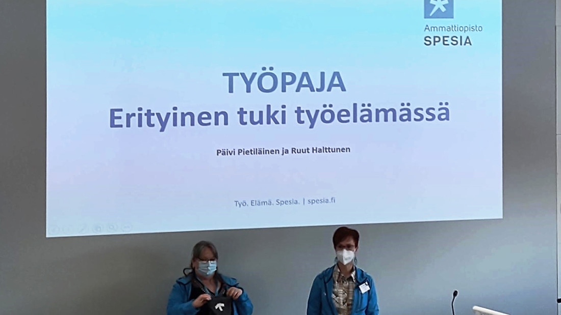 Esitystilanne auditoriossa, salin edessä seisoo kaksi naista Spesia-huppareissa ja takana näkyvällä dialla teksti "TYÖPAJA, Erityinen tuki työelämässä."