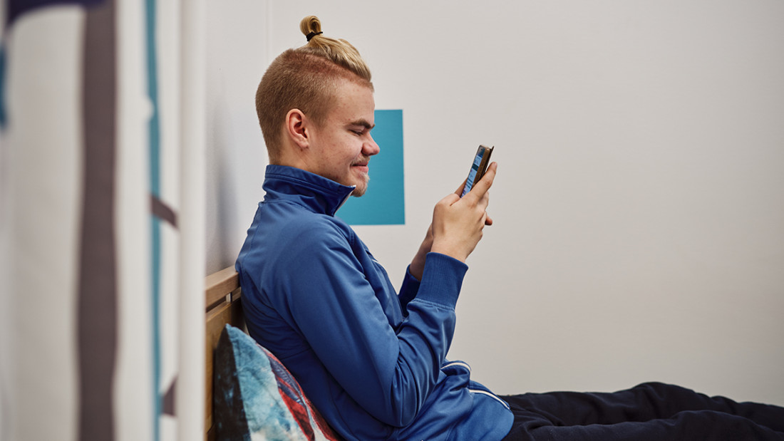Nuori mies istuu sängyllä ja katsoo kännykkää.