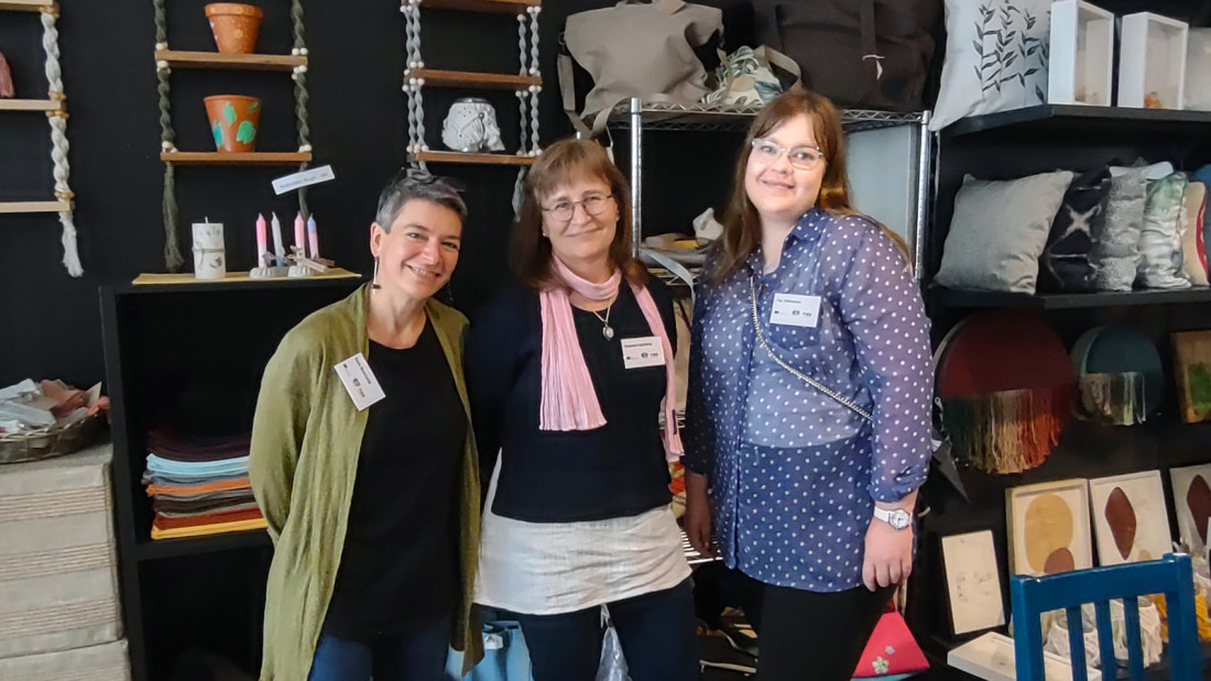 Kolme naista seisoo vierekkäin, esillä on erilaisia käsitöitä.