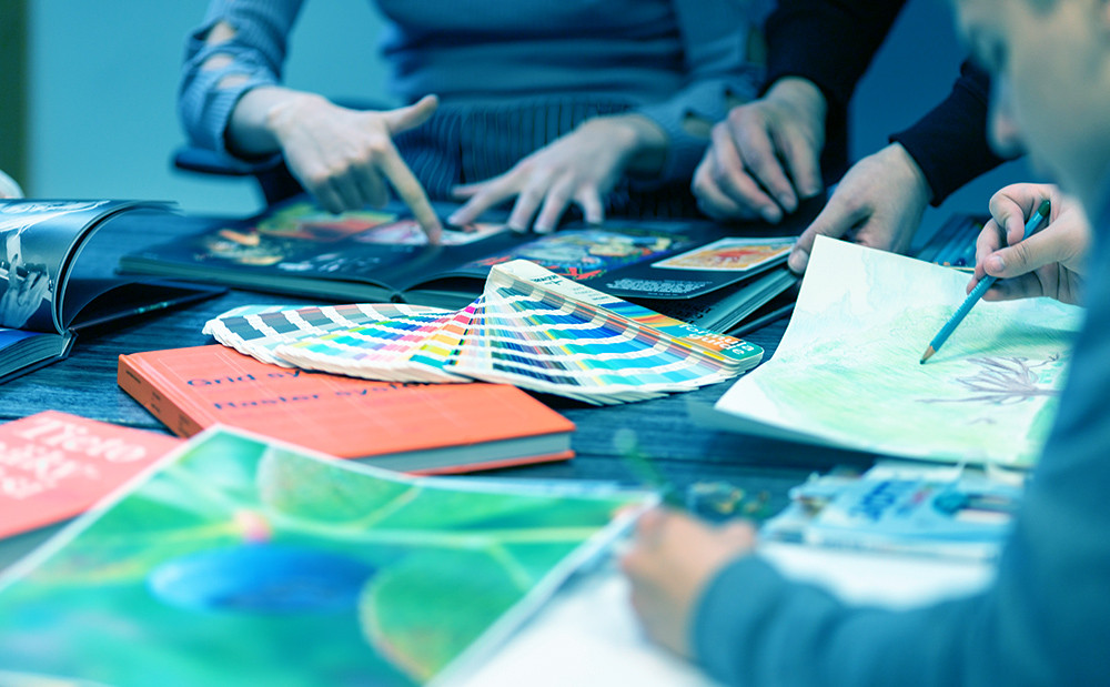 Pöydällä papereita, kirjoja ja väriviuhka, joiden lisäksi kuvassa näkyy kolmen pöydän ääressä istuvan henkilön kädet.