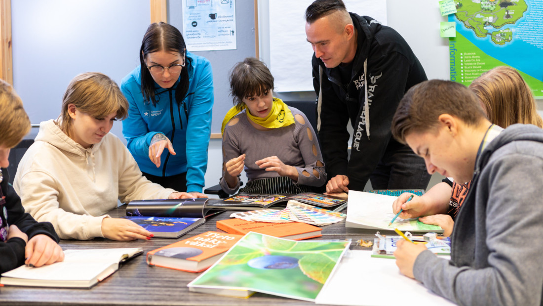 Opiskelijoita istuu pöydän ympärillä, vieressä seisoo opettaja ja ohjaaja, kaikki tutkivat materiaaleja pöydällä.