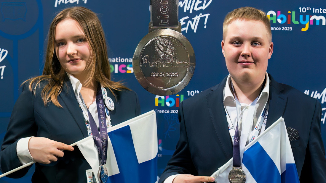 Nuori nainen ja mies Suomen liput käsissään, keskellä hopeamitali.