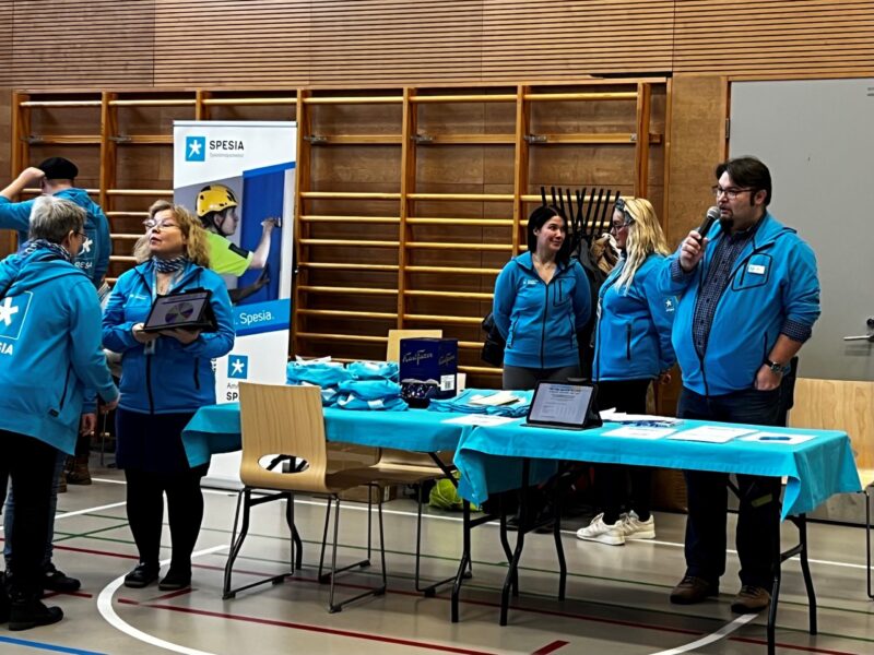 Liikutnasalissa esittelypöytä, spesian henkilöstöä ja mies mikrofoni kädessä.