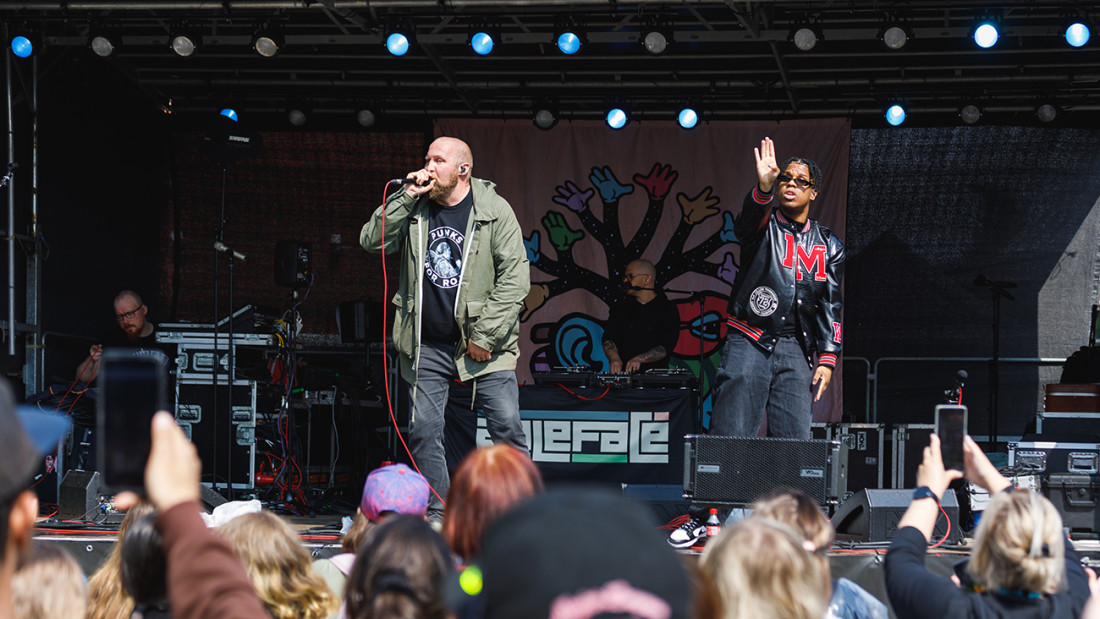 Konserttilavalla vihreään takkiin pukeutunut kalju mies laulaa ja mustaan takkiin pukeutunut mies lyhyissä rastahiuksissa viittoo.