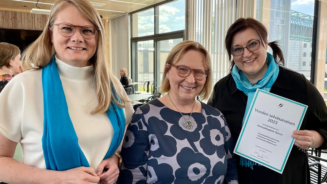 Kolme naista seisoo rinnakkain, katsoo kameraan ja hymyilee. Yksi pitelee kädessään kunniakirjaa.