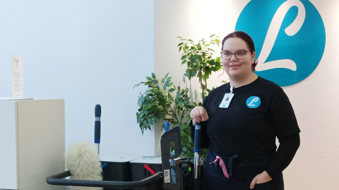 Nuori nainen seisoo siivouskärryn vieressä ja hymyilee hiema, taustalla seinässä näkyy Lounatuulen L-tunnus.