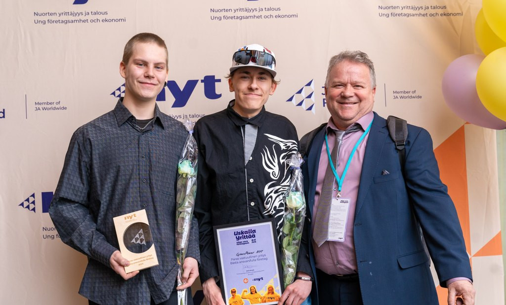 Kaksi opiskelijaa ja opettaja poseeraavat NYT-logoseinän edessä, opiskelijoilla on käsissään Uskalla Yrittää -kisan palkinnot.