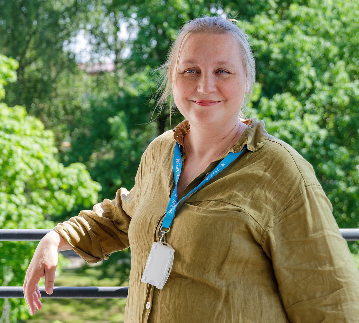 Nainen vihreässä paidassa seisoo nojaten parvekkeen kaiteeseen, taustalla kesäinen piha. Kaulassa on Spesian kaulanauha ja henkilökortti.