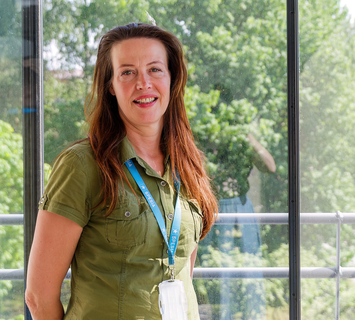 Nainen vihreässä paidassa seisoo ison ikkunan edessä, taustalla kesäinen maisema. Kaulassa on Spesian kaulanauha ja henkilökortti.