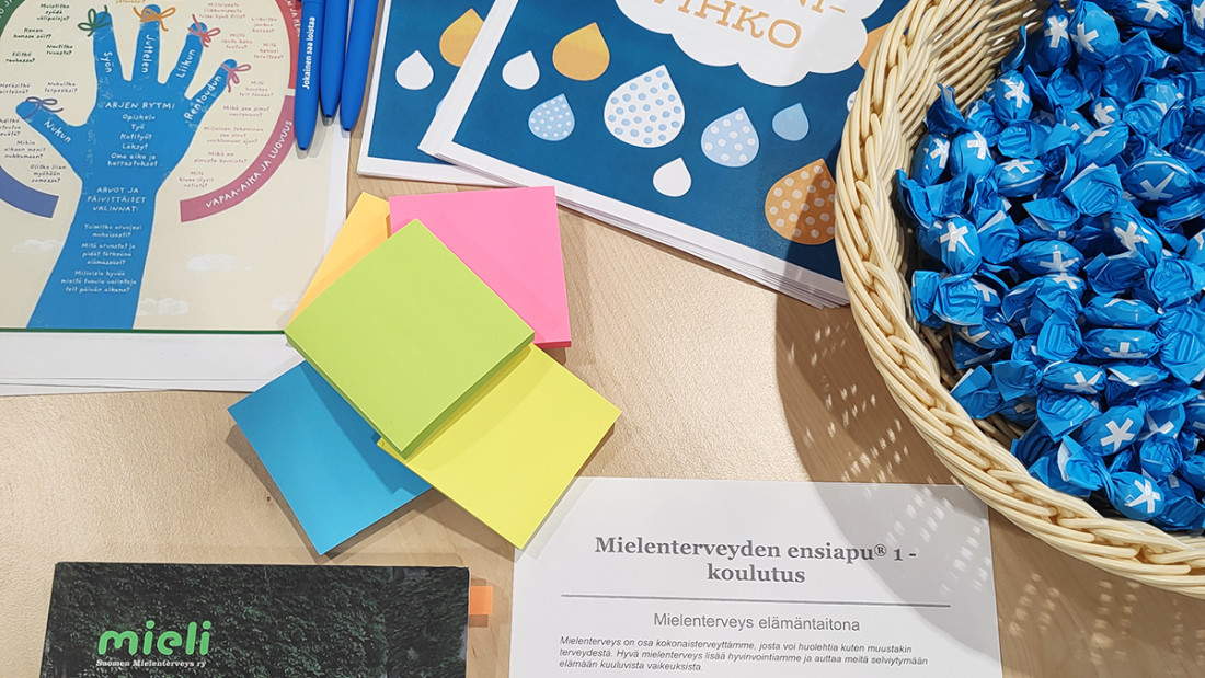 Mielenterveyteen liittyviä materiaaleja levällään pöydällä sekä muistilappuja ja korillinen Spesia-karkkeja.