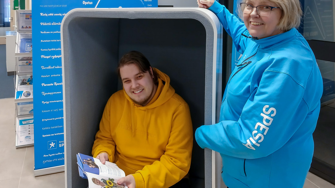 Spesian huppariin pukeutunut nainen seisoo, keltapaitainen nuori istuu hakijan opas kädessään.