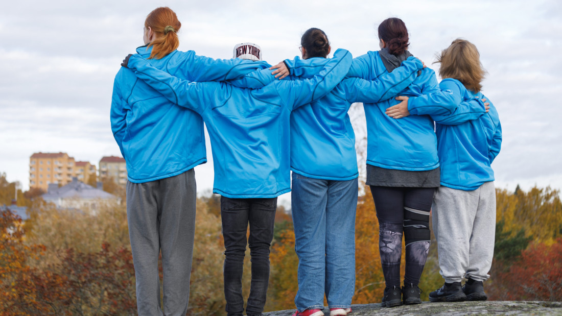 Viisi henkilöä seisoo sinisissä huppareissa vierekkäin selkämys kohti kameraan.
