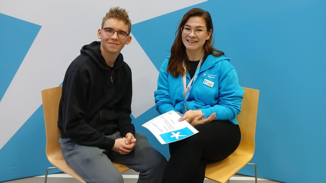 Miesopiskelija ja nainen Spesian sinisessä henkilöhupparissa istuvat tuoleilla.