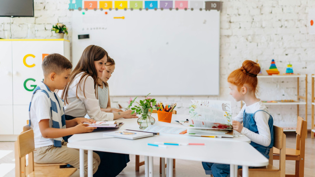 Kolme lasta ja yksi aikuinen istuvat luokkahuoneessa pöydän ympärillä, pöydällä on kirjoja ja kyniä.