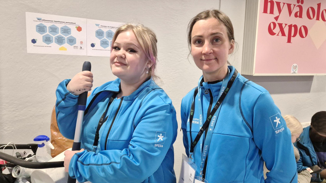 Kaksi Spesia-huppareihin pukeutunutta henkilöä, taustalla "hyvä olo expo" -teksti ja etualalla hieman siivousvälineistöä.