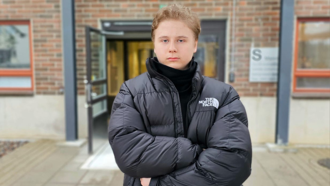 Nuori mies on pukeutunut mustaan takkiin ja seisoo kädet puuskassa tiilirakennuksen edessä.