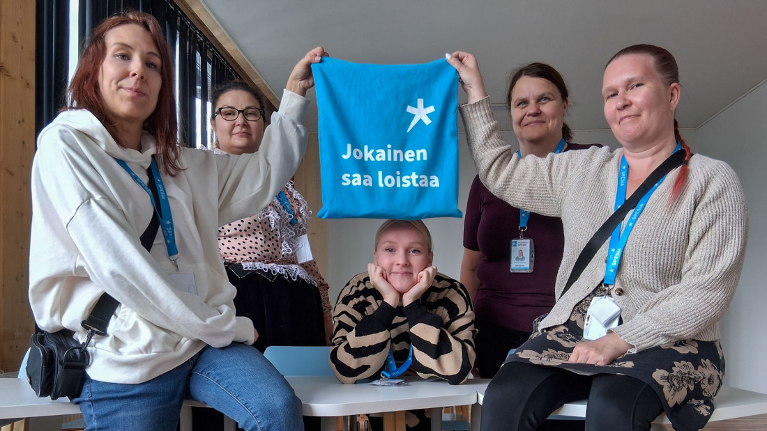 Viisi ihmistä pöydän ympärillä. Kaksi heistä pitelee välissään sinsitä kassia, jossa lukee "Jokainen saa loistaa" ja yksi kurkistaa sen alta nojaten leukaa käsiinsä.