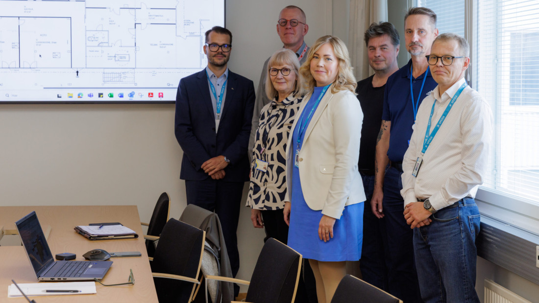 Kuusi henkilöä seisoo kokoushuoneessa suuren näytön edessä, jossa näkyy pohjapiirros. Etualalla kokouspöytä, jonka päällä on kannettava tietokone ja toimistotarvikkeita.