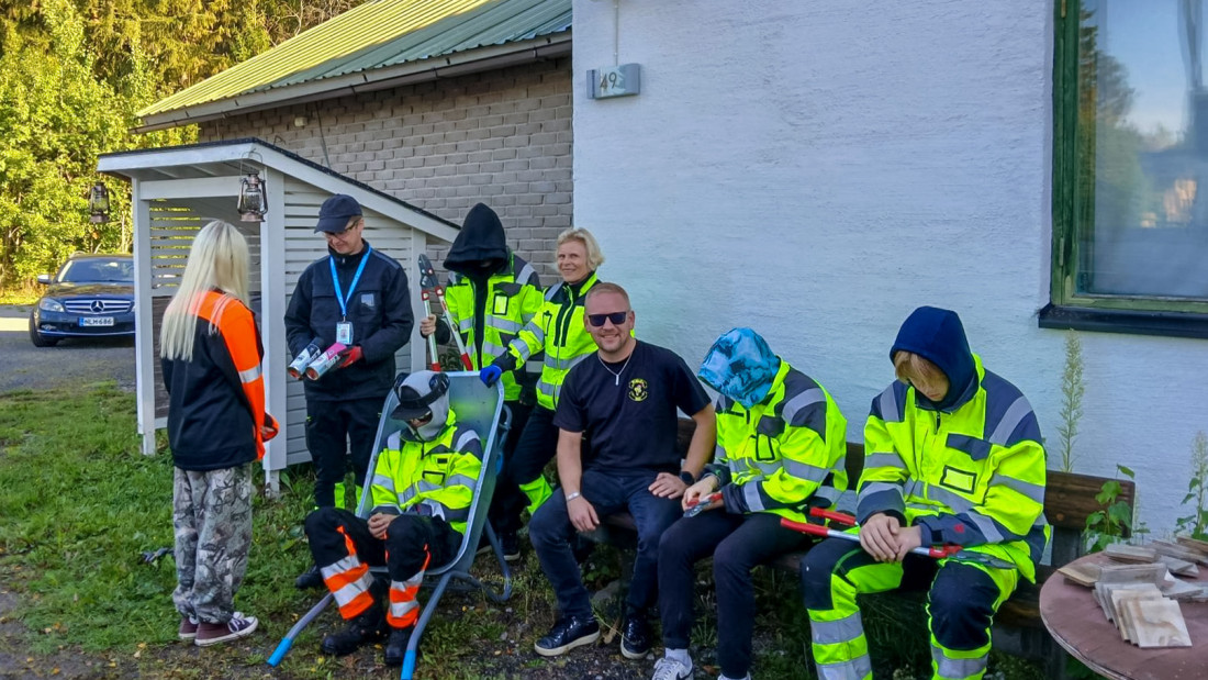 Joukko ihmisiä istuskelee talon seinustalla, suurin osa huomiovärisissä työvaatteissa. Ylhäällä MC PiMoKen kyltti.