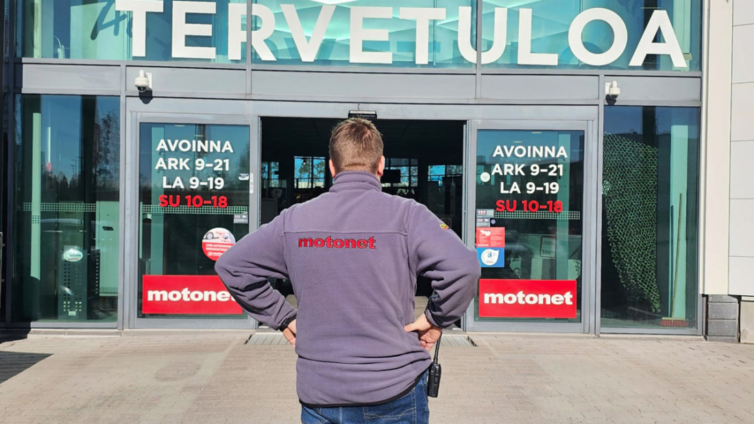 Mies seisoo kädet lanteilla ulkona, Motonetin oven edessä. Oven päällä lukee tervetuloa.