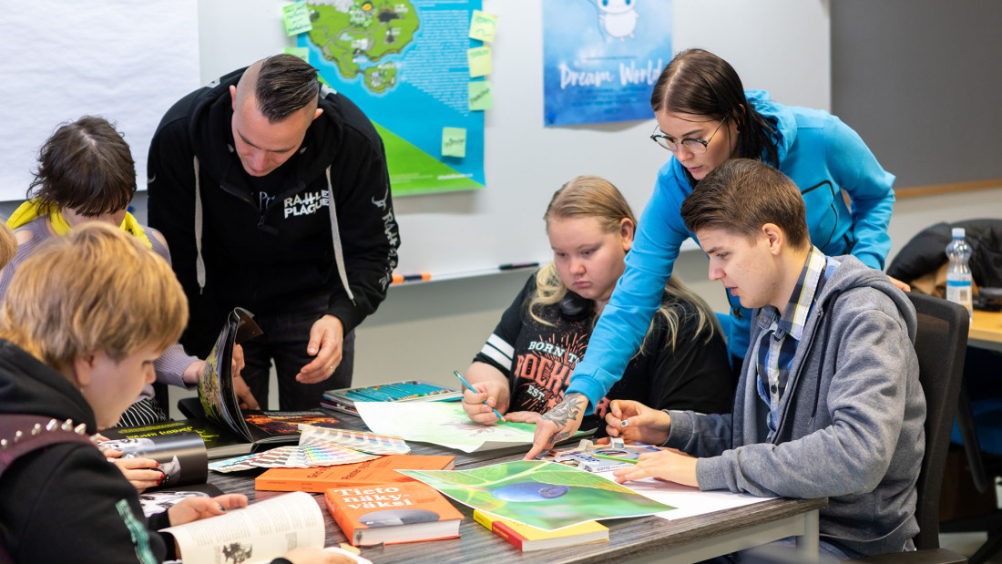 Kaksi henkilöä ohjaamassa pöydän ympärillä istuvia opiskelijoita luokkatilassa.