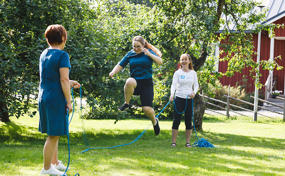 Kaksi henkilöä pyörittää hyppynarua kolmannen hyppiessä aurinkoisena kesäpäivänä.
