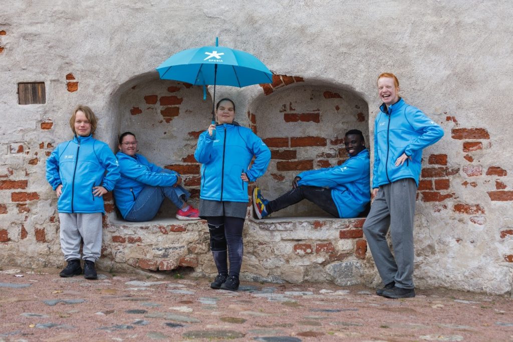 Spesian opiskelijoita Spesia-hupparit päällään kiviseinän vieressä. Yhdessä opiskelijalla on sateenvarjo kädessään.