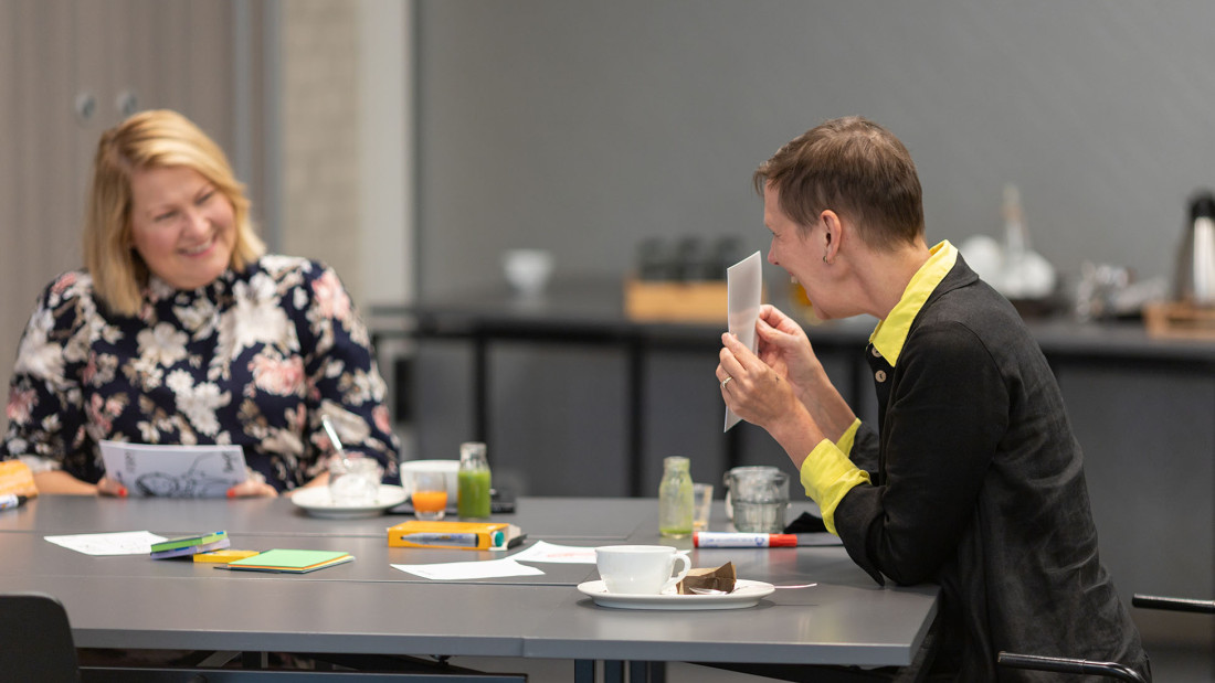 Kaksi nauravaa henkilöä istuu pöydän äärellä, toinen näyttää paperia toiselle.