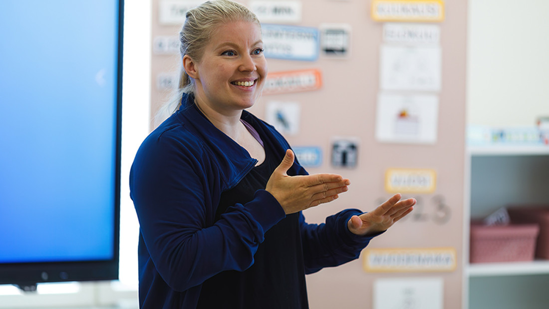 Iloinen nainen seisoo luokkahuoneessa ja kommunikoi käsillään.