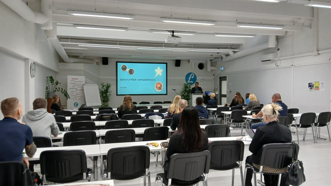 Luentosali, jossa istuu ihmisiä tuoleilla katsoen edessä olevaa näyttöä, jolla Spesian esitys.