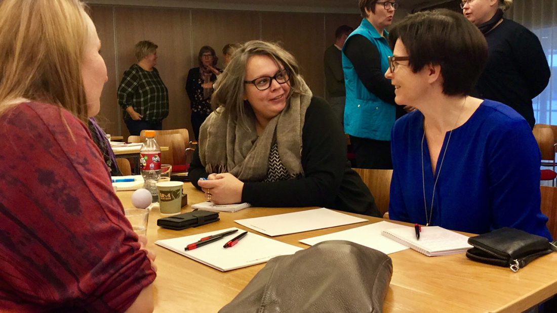 Kolme henkilöä keskustelevat pöydän äärellä ja taustalla muita henkilöitä.