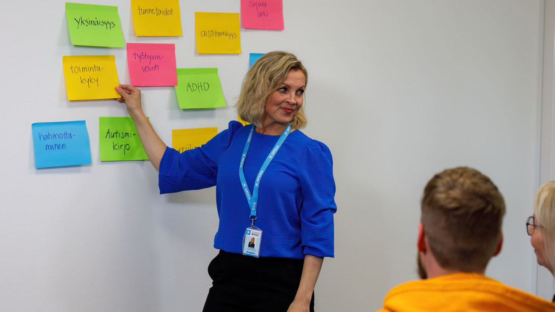 Seinällä on nepsy-aiheisia sanoja sisältäviä erivärisiä liimalappuja, kouluttaja Tuula Niskanen osoittaa niistä yhtä kahdelle kuuntelevalle henkilölle.