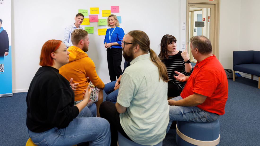 Kaksi kouluttajaa seisoo taka-alalla, etualalla on ihmisiä istumassa ja keskustelemassa pareittain.