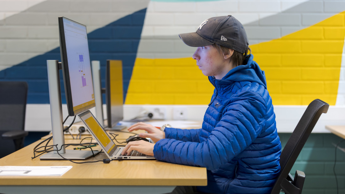 Opiskelija tietokoneen äärellä, hänellä on käytössään sekä läppäri että iso näyttö.