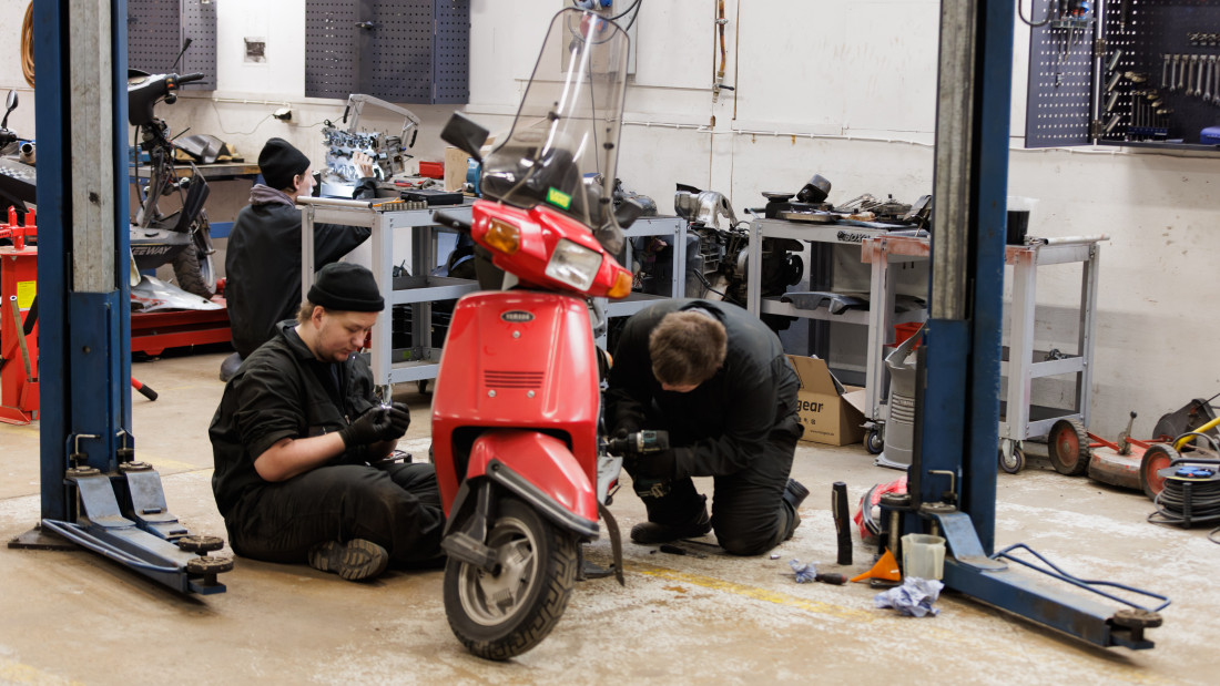 Opiskelijat työntouhussa korjaamolla, edessä punainen skootteri.