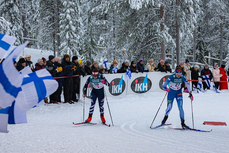 Hiihtäjiä kisaladulla ja katsojia kannustamassa aidan takana.