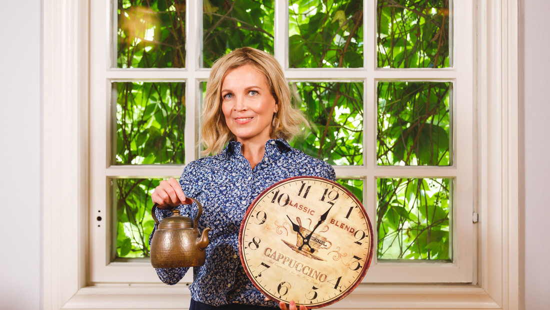 Nainen seisoo selin ikkunaan ja pitää toisessa kädessään kuparista kahvipannua ja toisessa kädessä seinäkelloa.