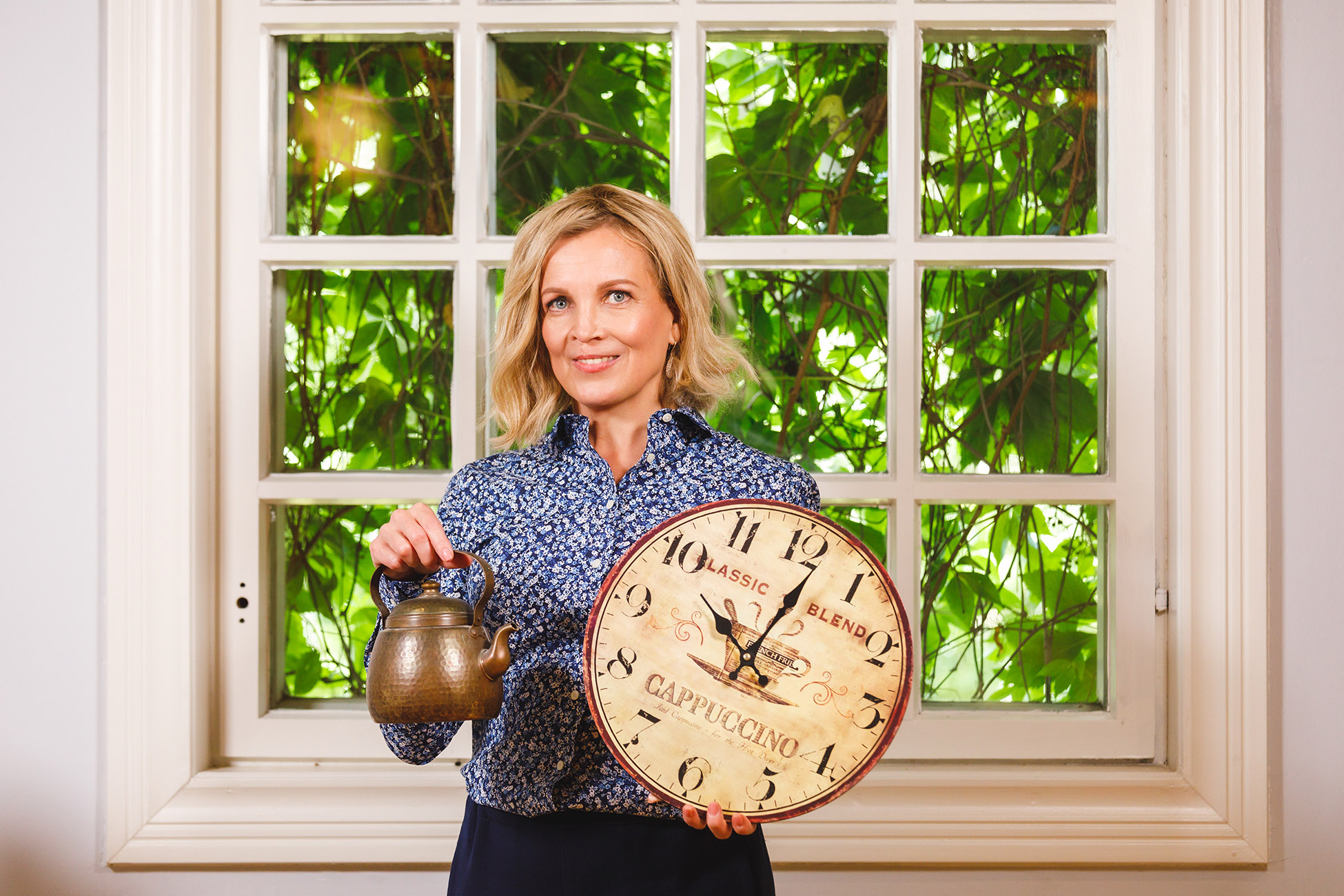 Nainen seisoo selin ikkunaan ja pitää toisessa kädessään kuparista kahvipannua ja toisessa kädessä seinäkelloa.