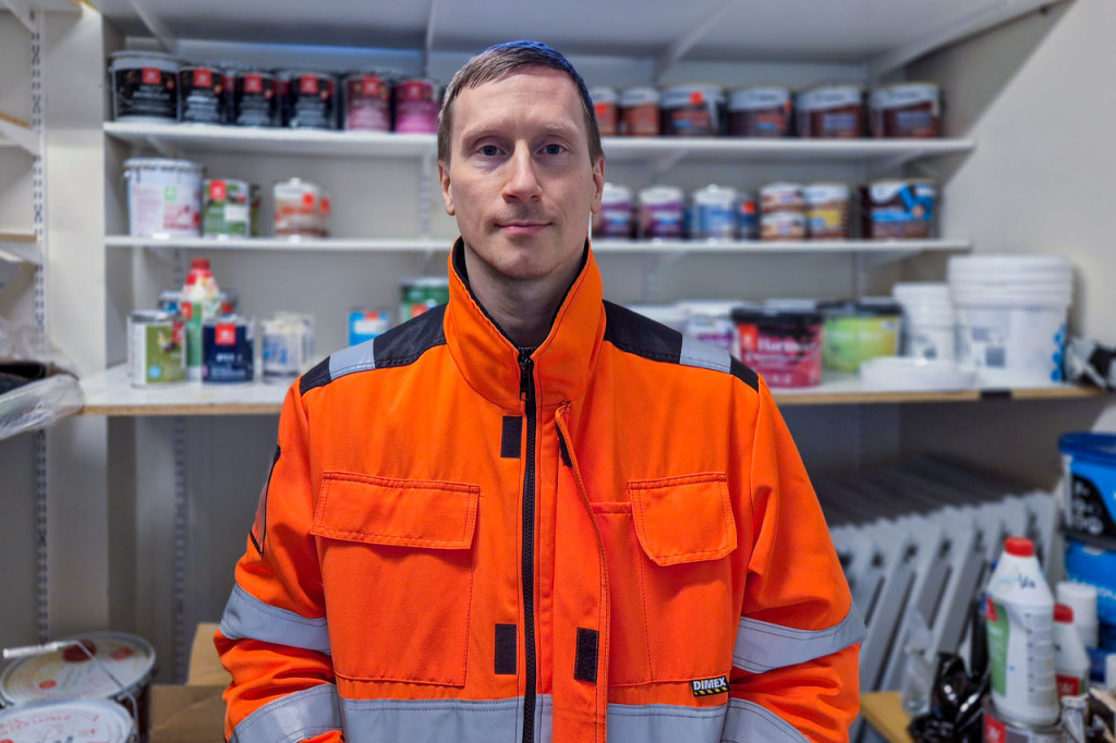 Mies oranssissa työtakissa seisoo varastohuoneessa ja katsoo suoraan kameraan.