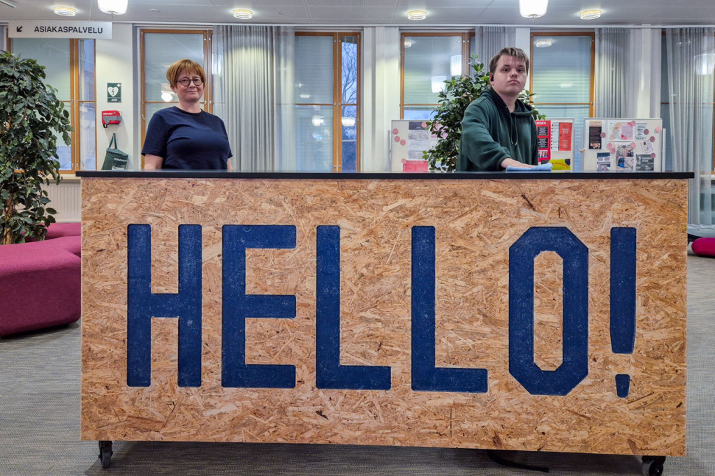 Opiskelija pyyhkii tiskiä oppilaitoksen tiloissa, aikuinen seuraa vieressä. Tiskissä lukee isosti Hello!