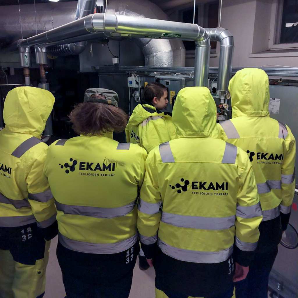 Keltaisiin työtakkeihin pukeutuneet opiskelijat seuraavat teknisessä tilassa kun yksi henkilö tekee jotain putkien luona. Takeissa lukee Esedu.