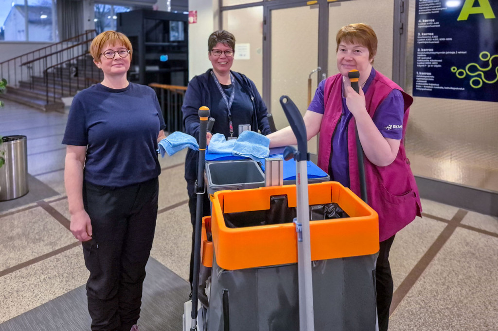 Kolme työasuihin pukeutunutta naista oppilaitoksen aulassa siivouskärryn ympärillä.