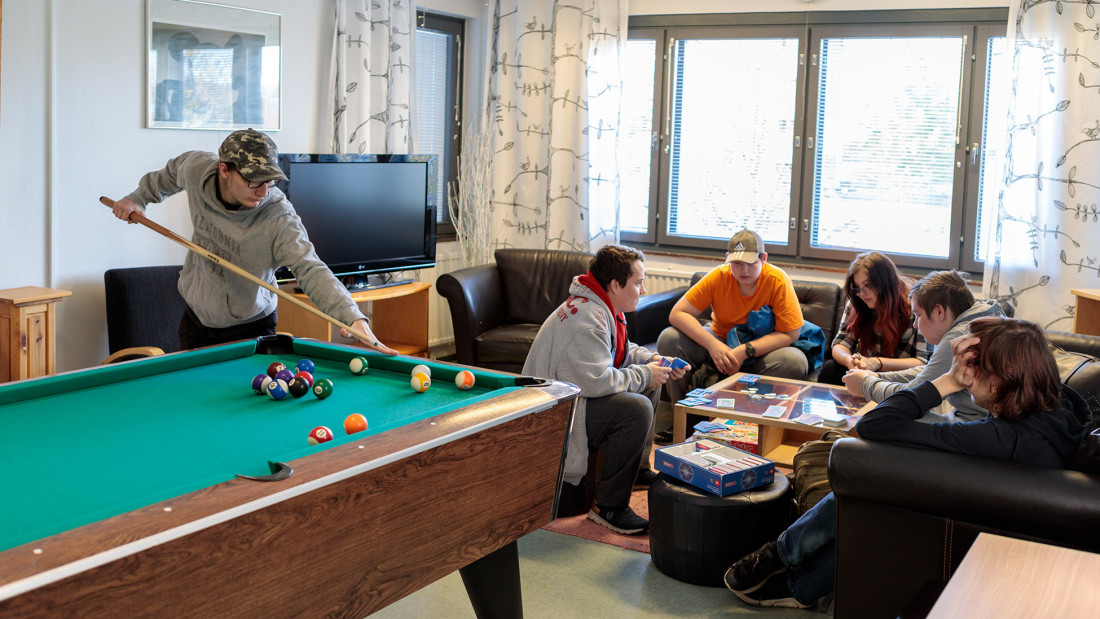 Opiskelijoita asuntolassa, yksi pelaa biljardia ja muut istuvat sohvalla pelaamassa.