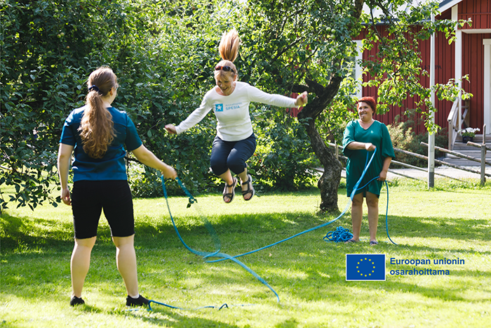 Kaksi henkilöä pyörittää hyppynarua ja yksi hyppii kesäisessä pihapiirissä, alalaidassa EU-lippulogo Euroopan unionin osarahoittama -tekstillä.